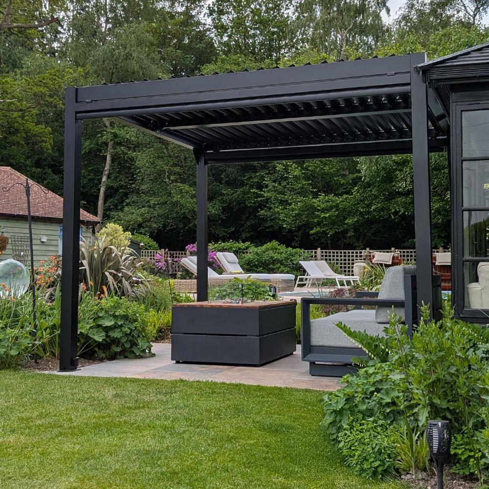 Fire table and seating under a pergola with a garden and pool visible beyond